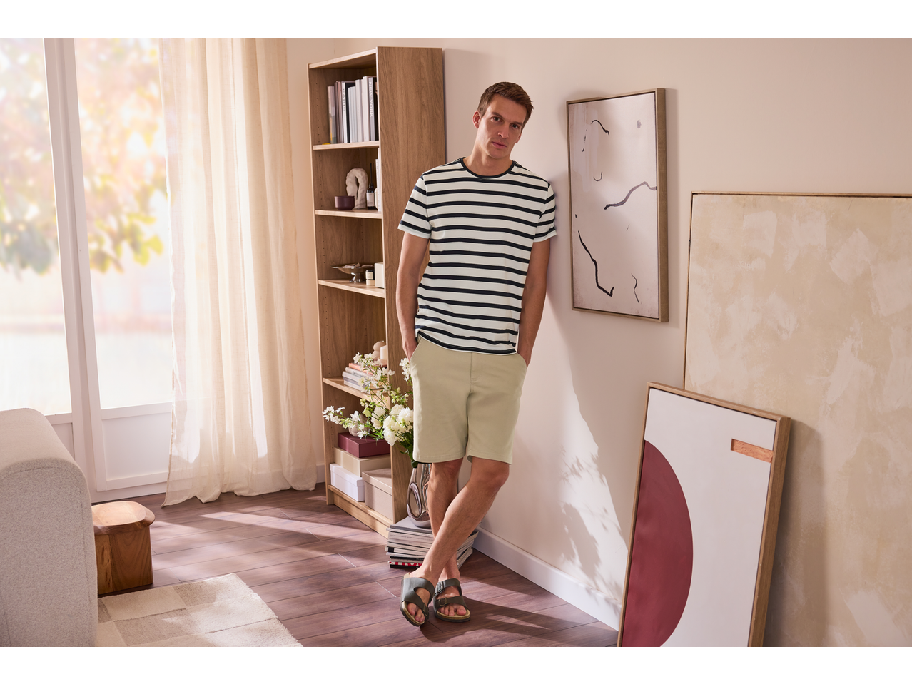 Muškarac u prugastoj majici i kaki šortsu, naslonjen na zid sa apstraktnom umetnošću.