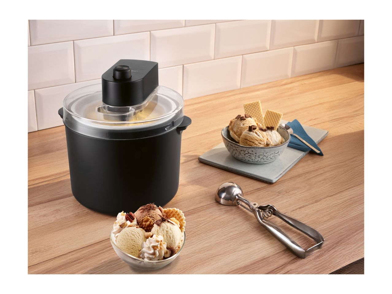 Ice cream maker on a wooden counter with bowls of ice cream and an ice cream scoop.