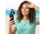 Mulher usando um ventilador de telefone na praia, com o oceano azul ao fundo.