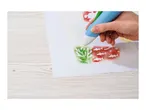 A hand using an airbrush pen to stencil an underwater scene with green coral and red fish.