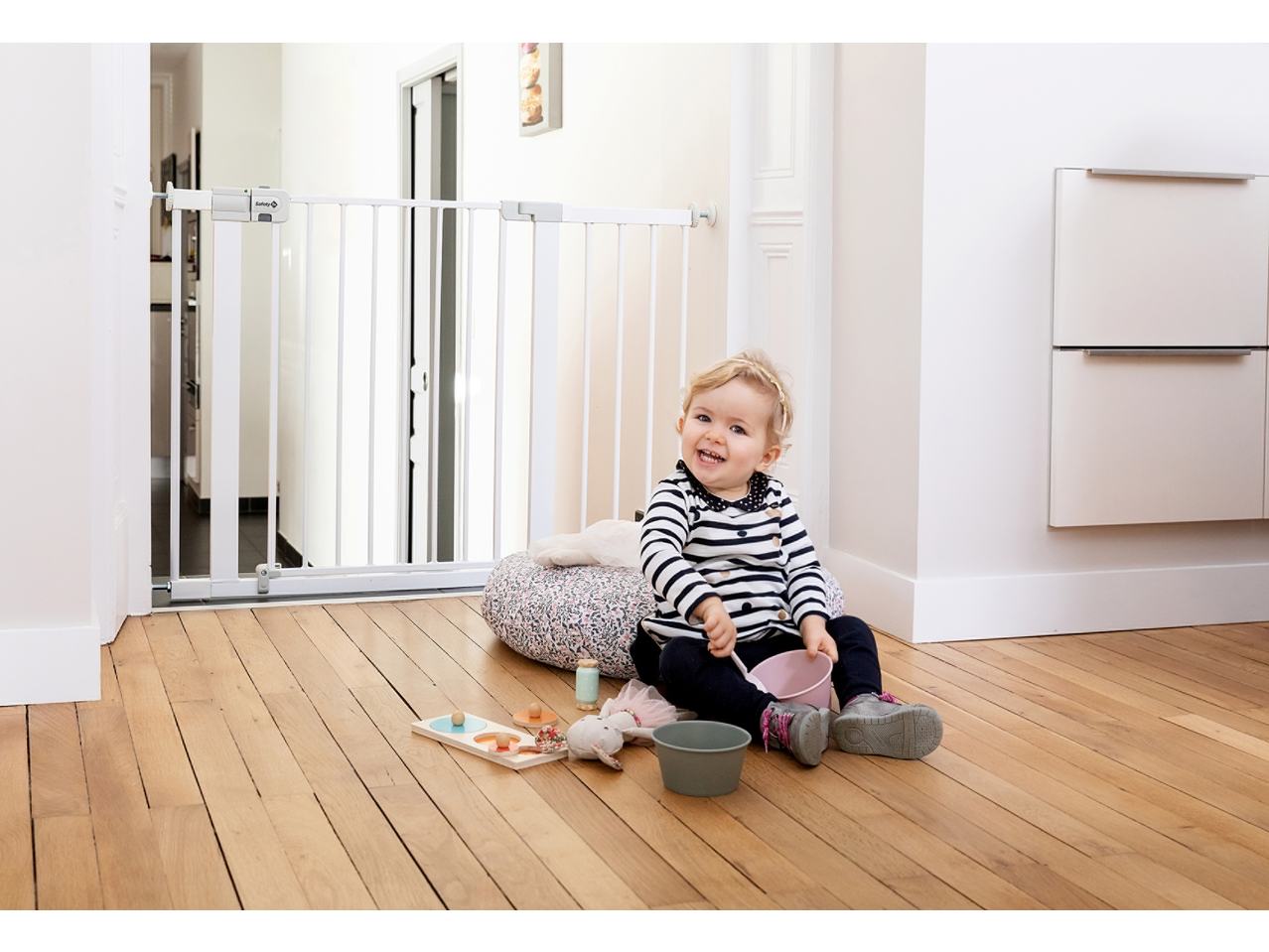 A baby sitting on a wooden floor, playing with toys, next to a white safety gate.