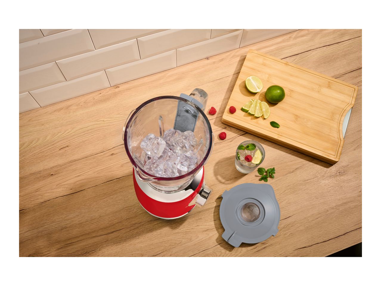 Red blender filled with ice cubes on a wooden countertop next to fresh limes and raspberries.