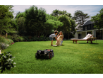 Robotic lawnmower on a green lawn with a family and dog in the background.