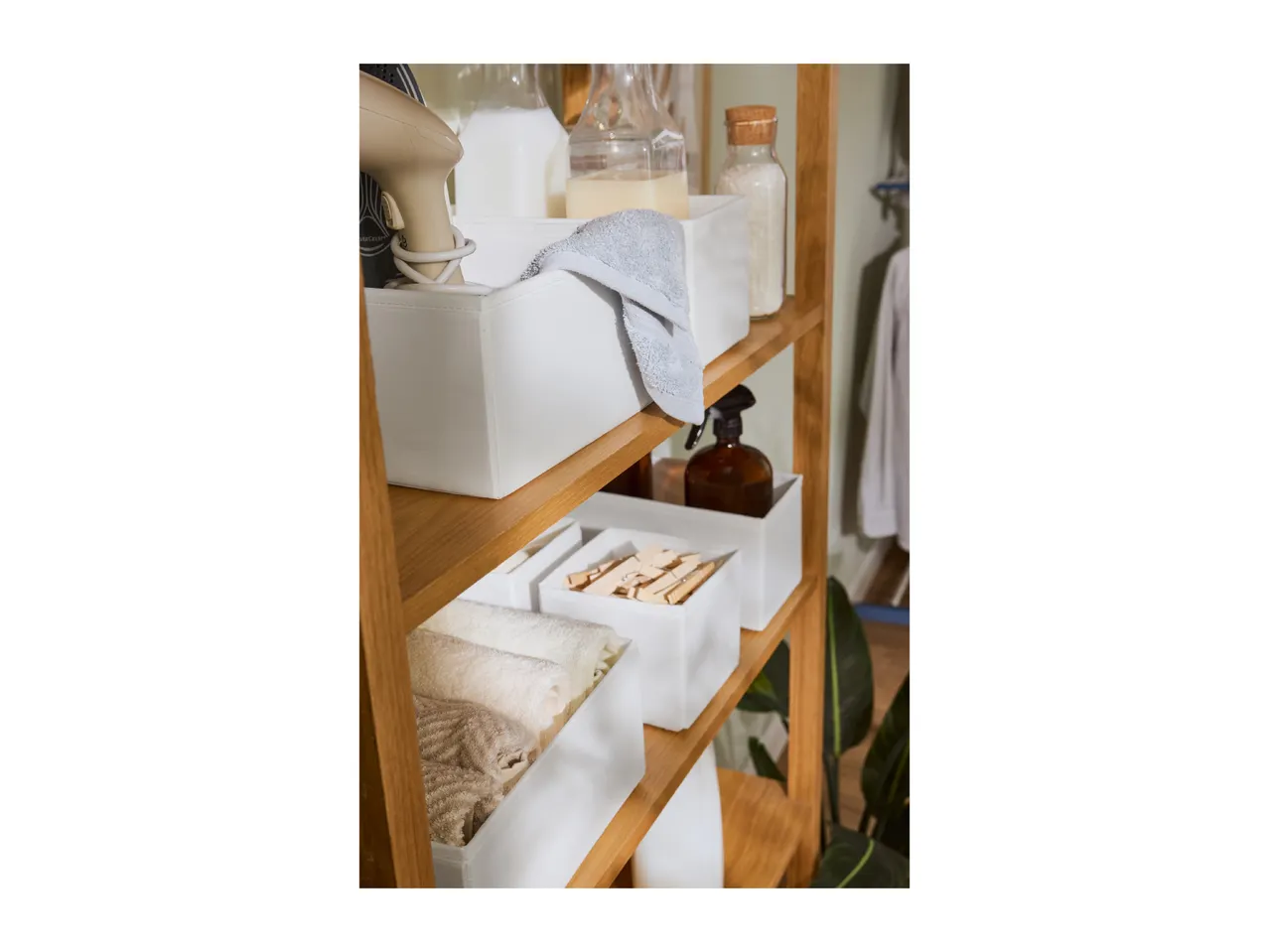 Wooden shelving unit with white storage boxes, containing laundry essentials like towels, clothes pegs, and liquid detergents.