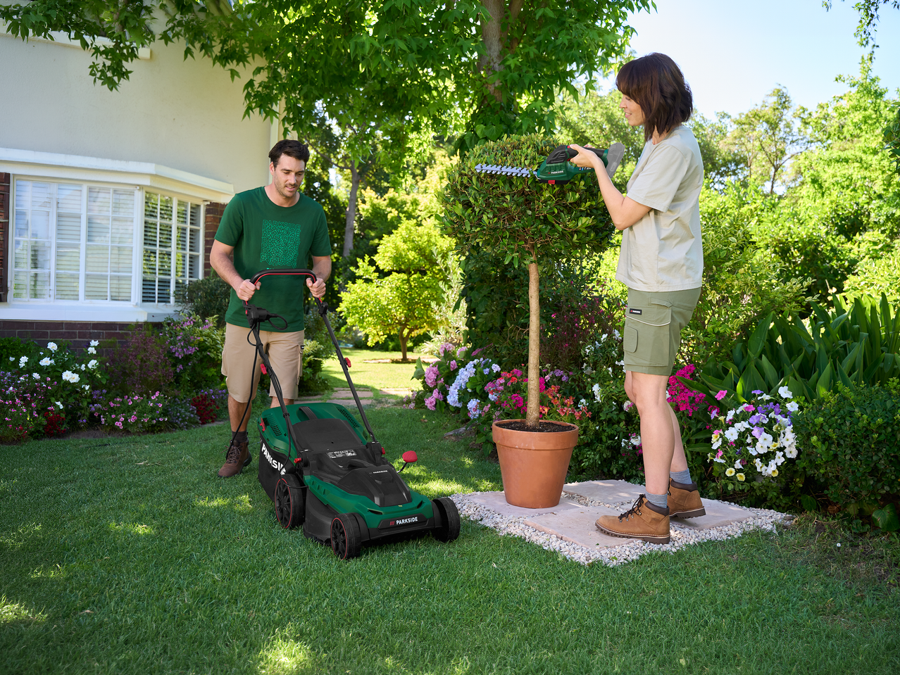 Άνδρας κουρεύει γκαζόν, γυναίκα κλαδεύει φυτό με ψαλίδι μπορντούρας σε ηλιόλουστο κήπο.
