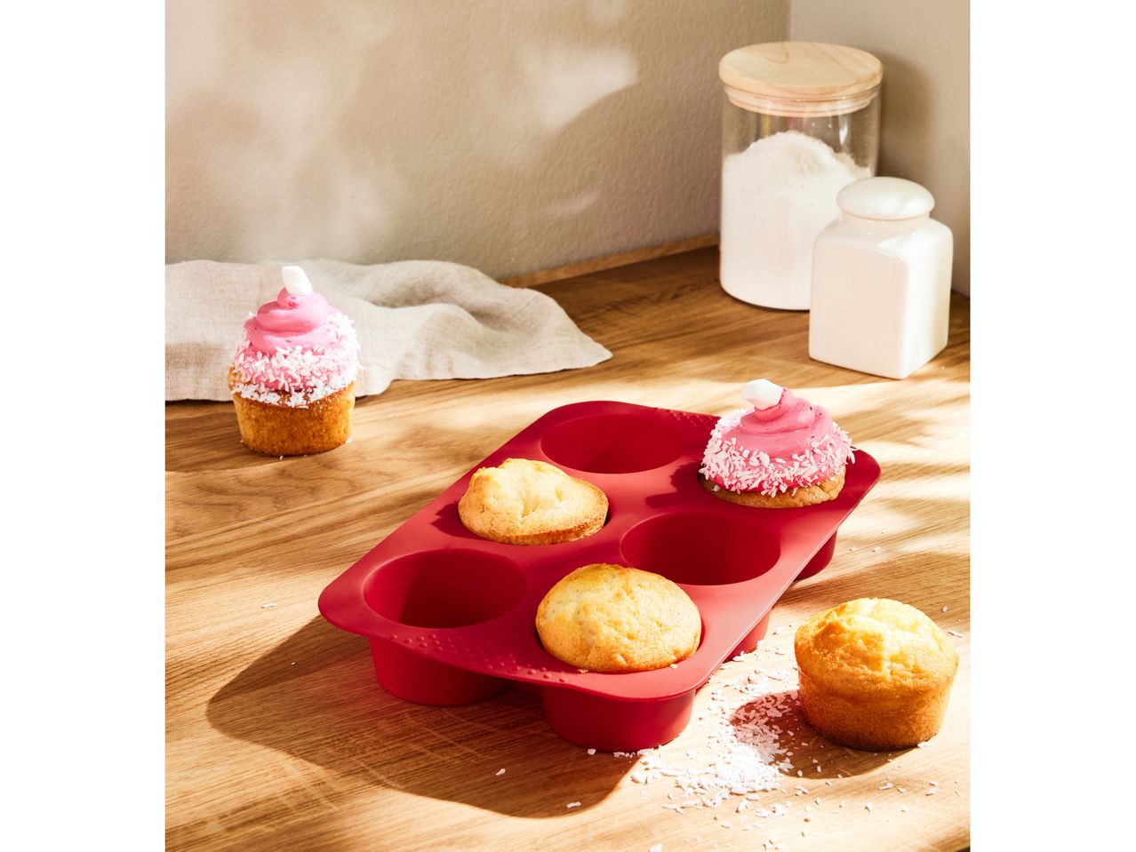 A red silicone muffin pan with baked muffins, one decorated with pink frosting and coconut flakes.