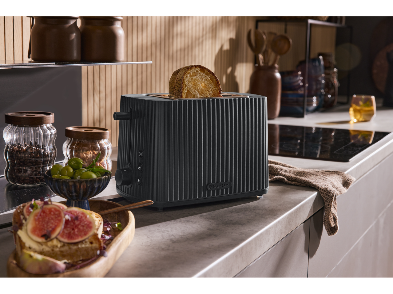 Prăjitor de pâine negru cu pâine prăjită într-o bucătărie, lângă boabe de cafea și măsline.