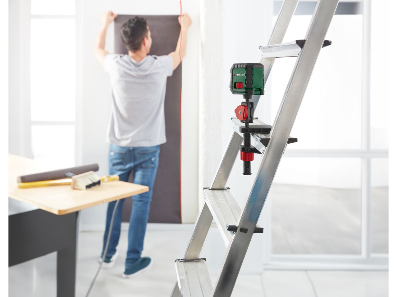 A Parkside laser level is mounted on a ladder, with a person in the background hanging wallpaper.