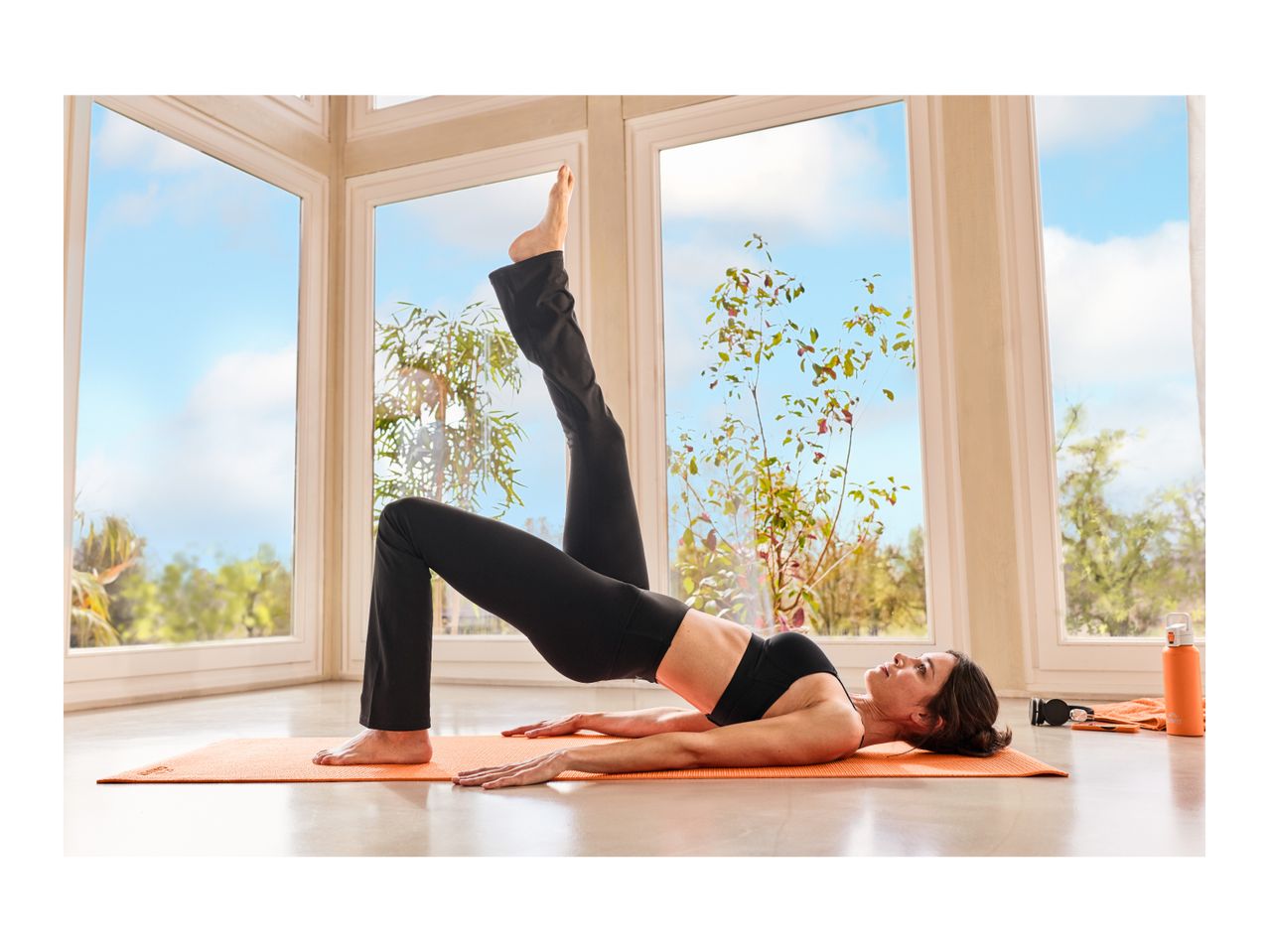 Woman in black activewear doing yoga on an orange mat by a window.