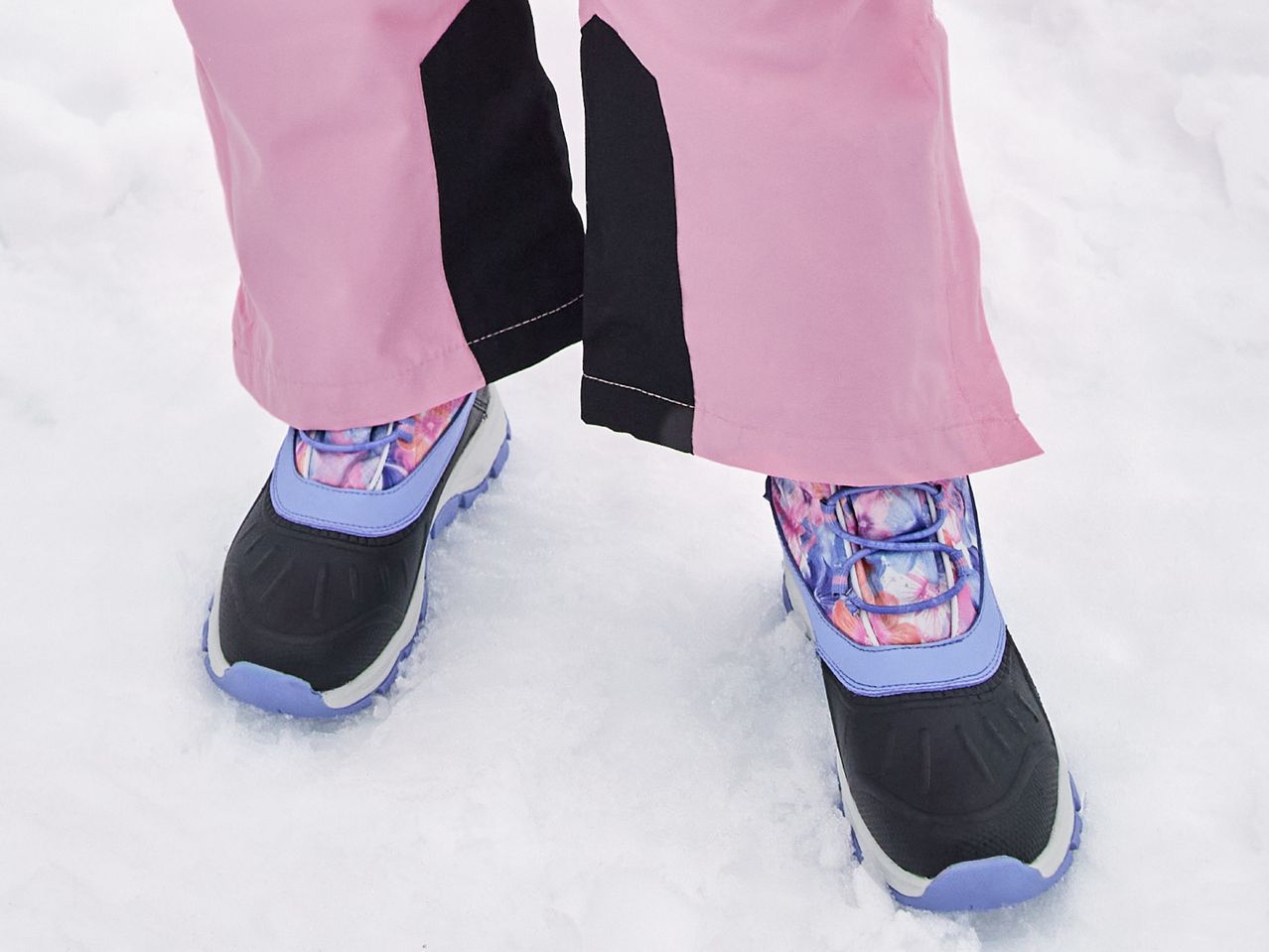Close-up of a person wearing pink snow pants and black and purple winter boots in snow.