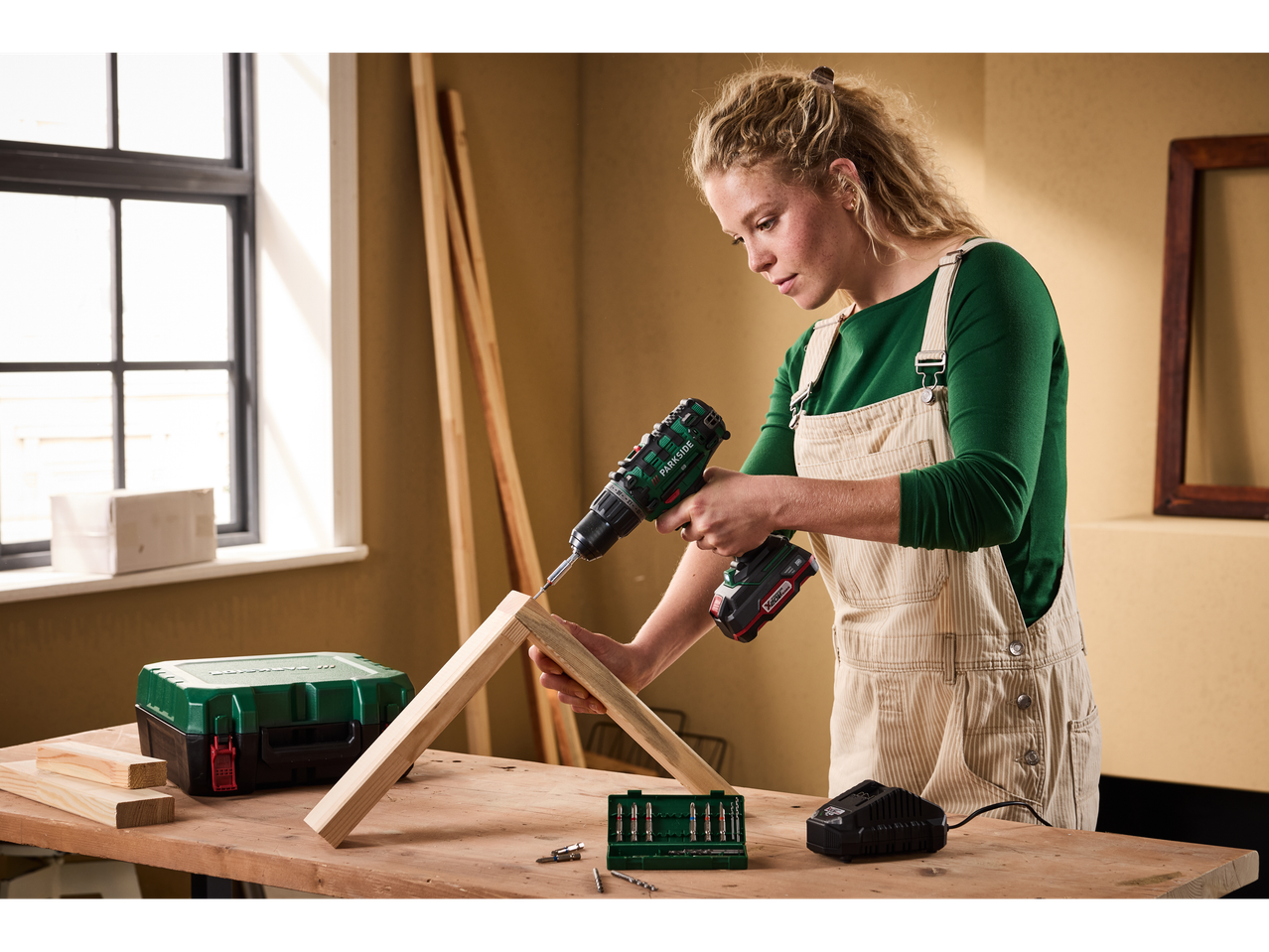 A woman uses a Parkside® 20V Cordless Drill Driver to assemble wooden pieces on a workbench.