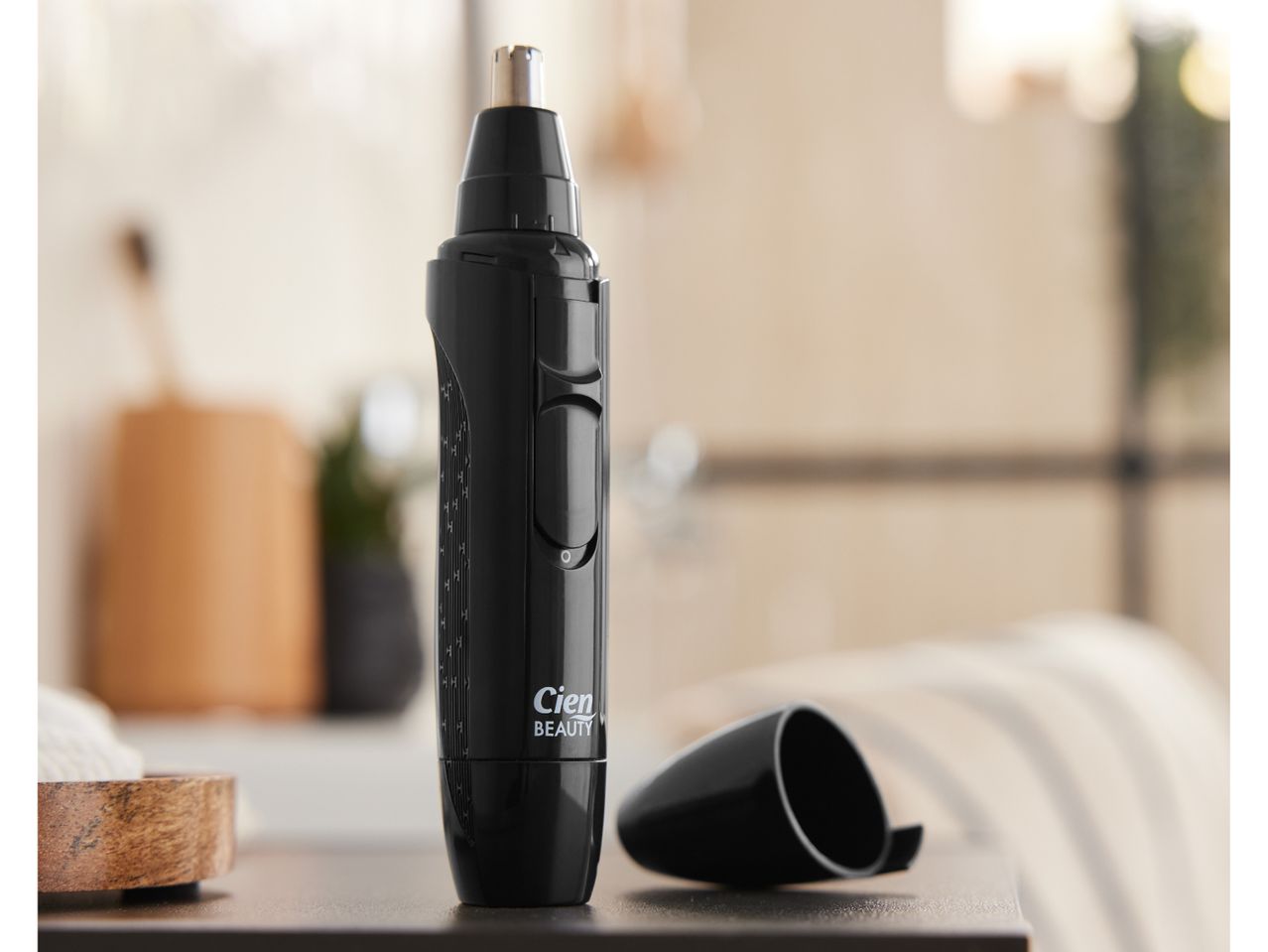 A black, cordless nose and ear hair trimmer sits on a table next to its protective cap.