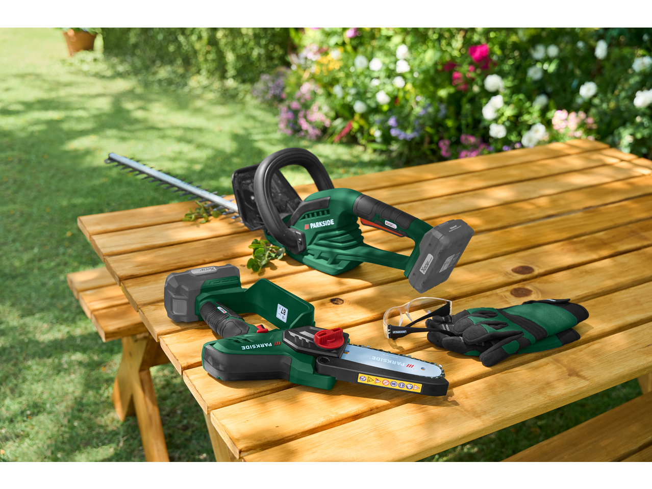 Aparador de sebes e motosserra a bateria PARKSIDE, com óculos de segurança e luvas, numa mesa de madeira.