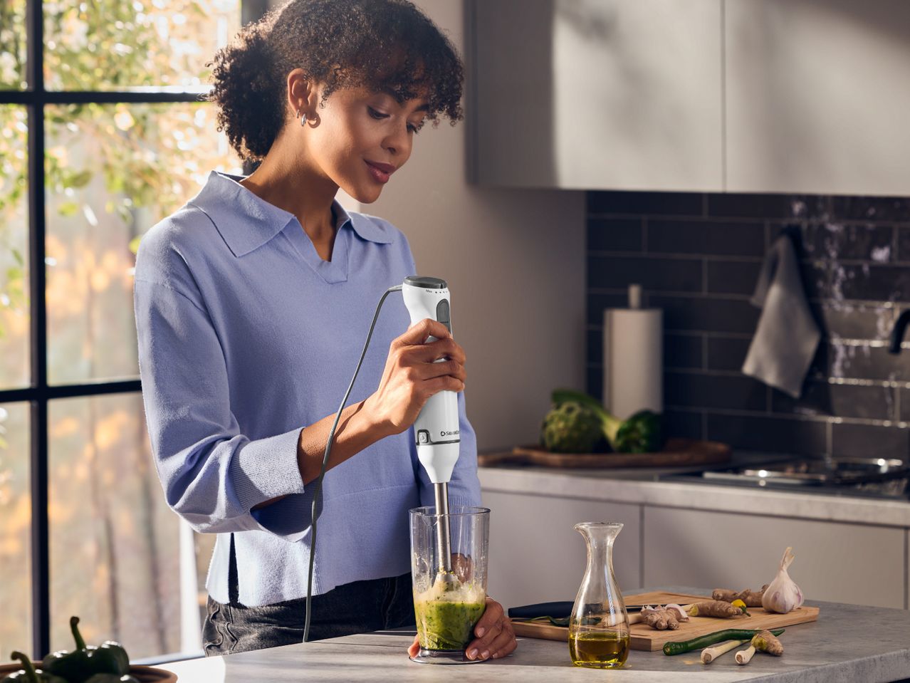 Woman blending green ingredients in a glass with an immersion blender in a kitchen.