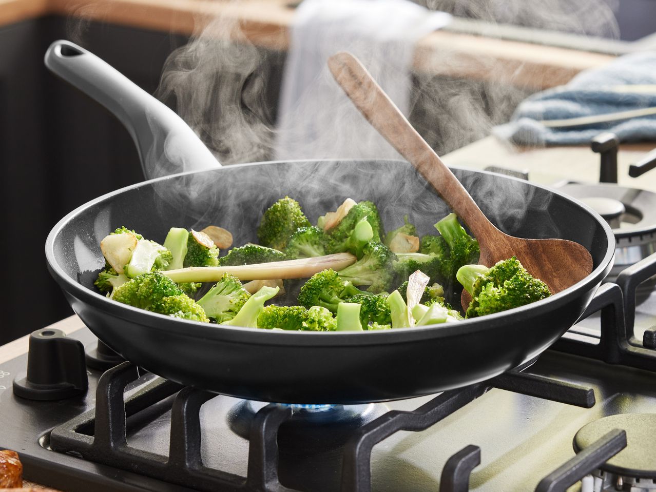 Steaming broccoli and other vegetables stir-fry in a dark-colored frying pan on a gas stove.