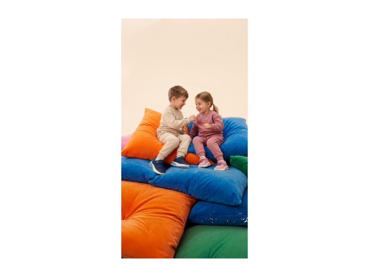 Two children in tracksuits sitting on colorful cushions, smiling and playing.