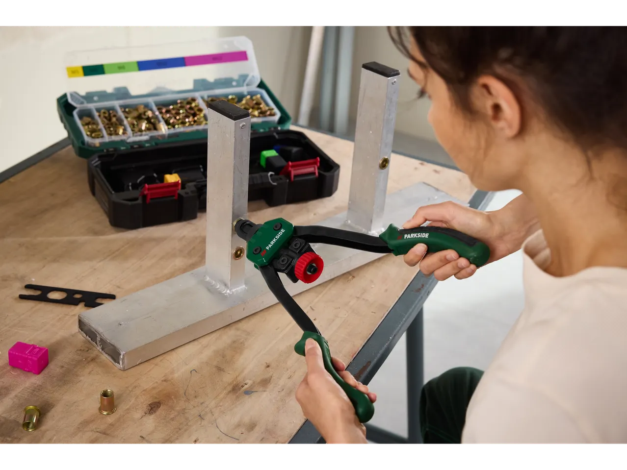 A person uses a rivet nut tool to attach a nut to an aluminum beam, with accessories nearby.