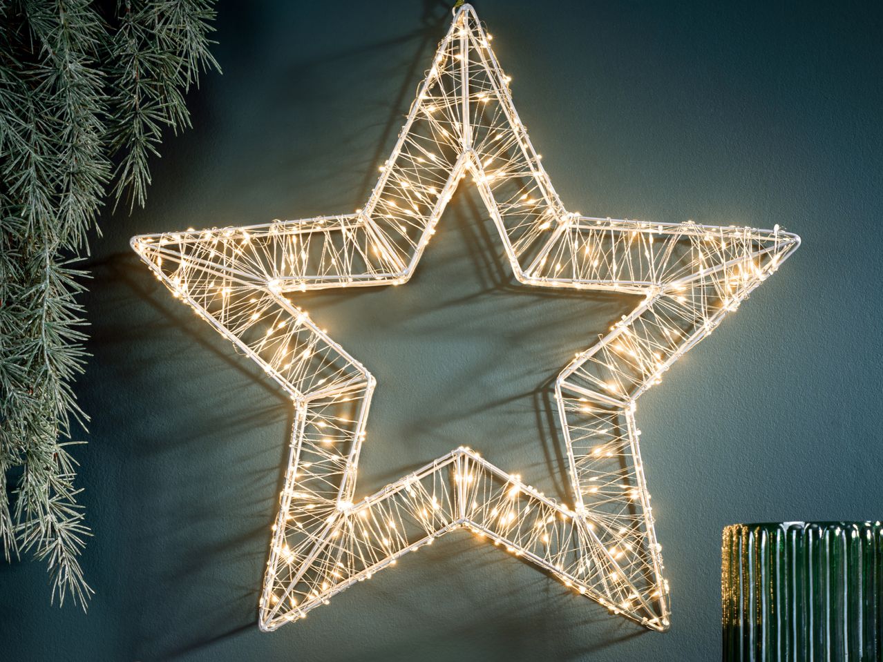 Светеща коледна звезда на тъмносиня стена с клонки от ела и зелена ваза.