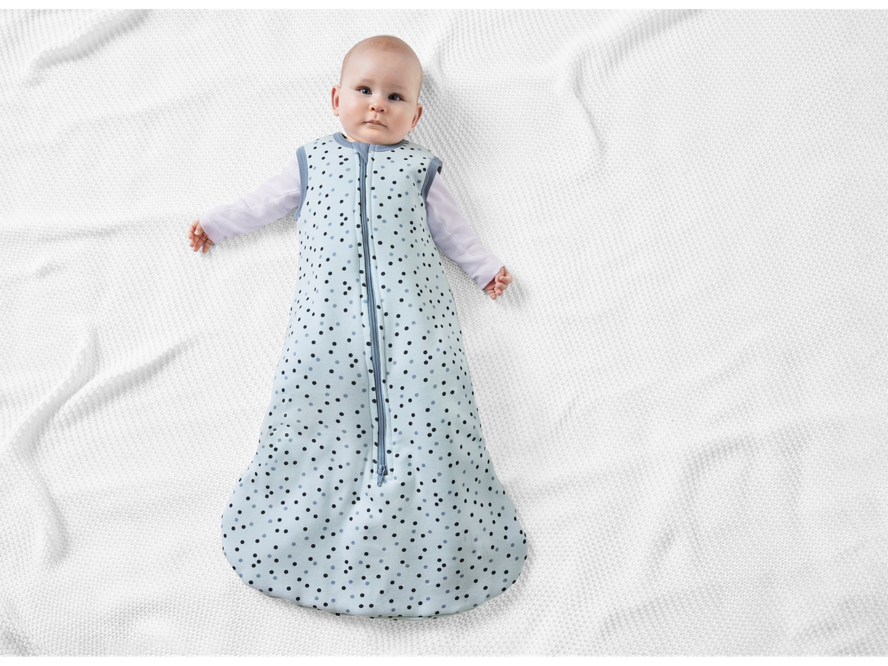 Baby in a light blue polka dot sleep sack on a white blanket.