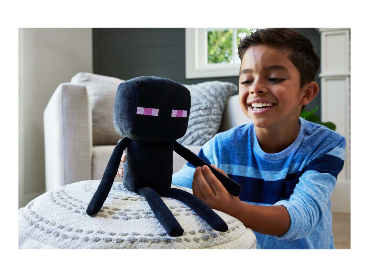 A boy playing with a black plush toy with pink eyes, sitting on a white knitted pouf.