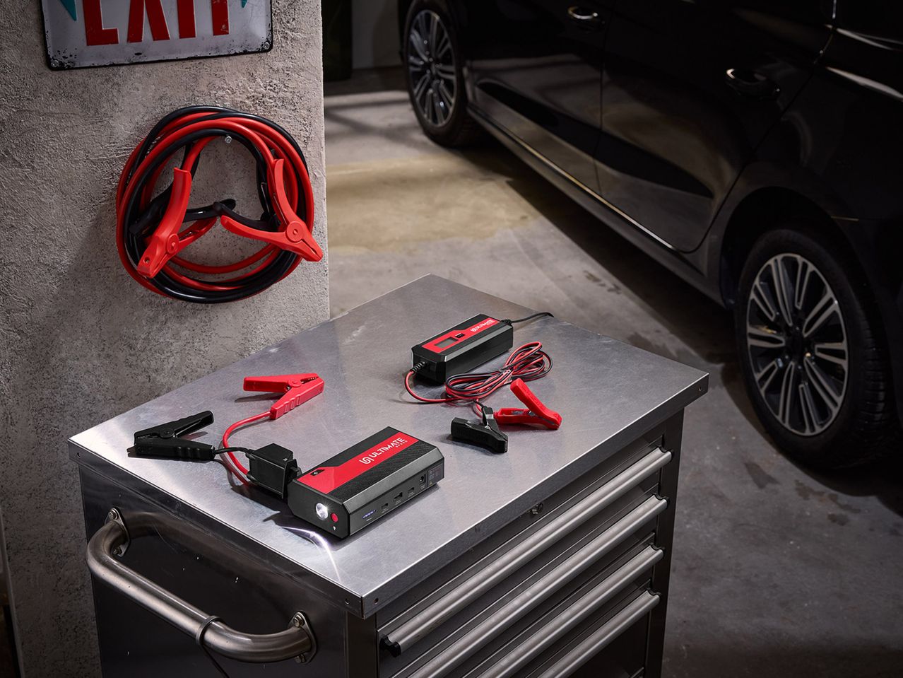 Εκκινητής μπαταρίας και φορτιστής μπαταρίας σε μεταλλικό τραπέζι εργαλείων με καλώδια εκκίνησης και αυτοκίνητο στο παρασκήνιο.