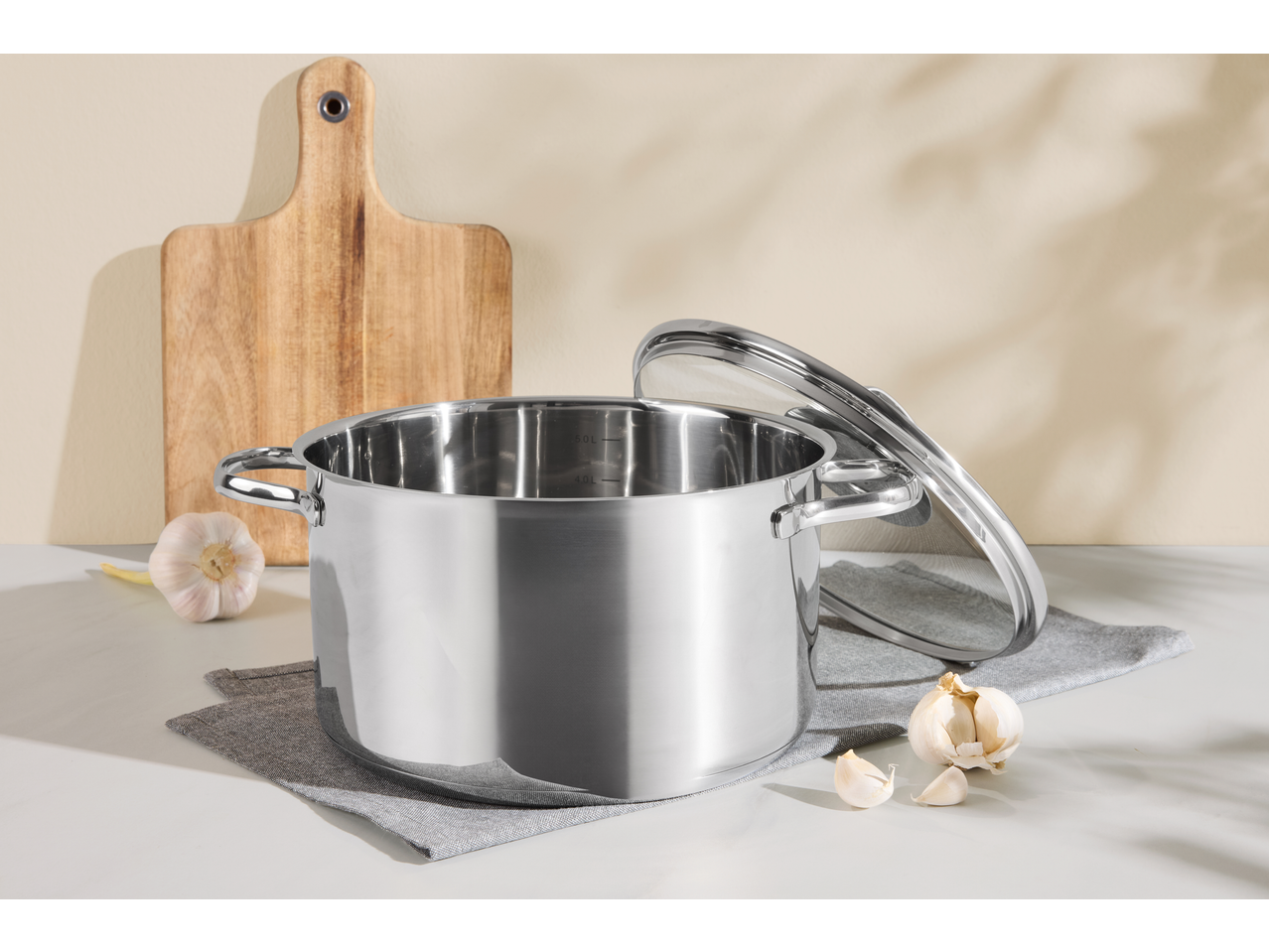 O oală de inox cu capac de sticlă, usturoi și un tocător de lemn pe un blat de bucătărie.