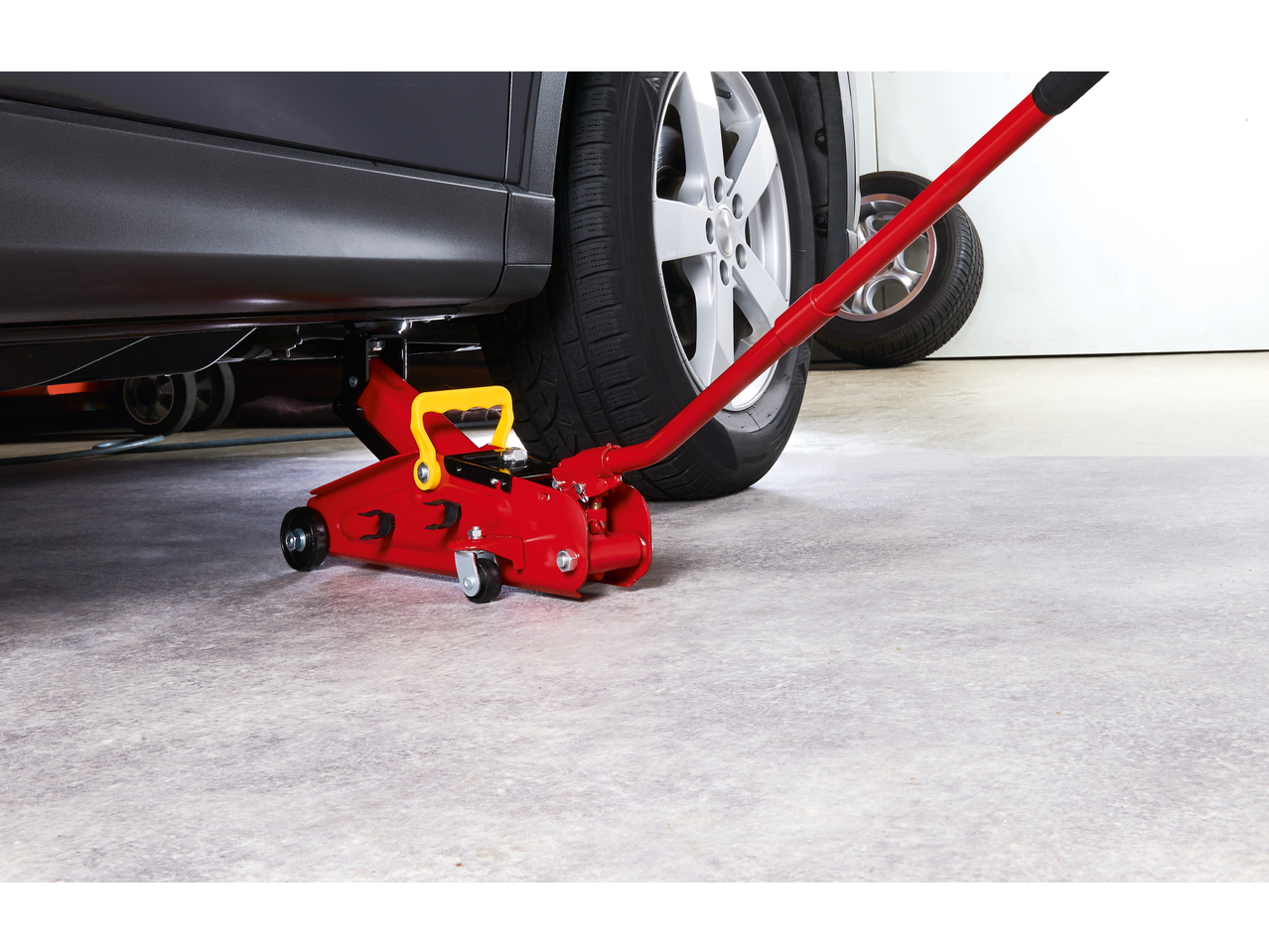 Red hydraulic car jack lifting a dark grey car, with a spare tire in the background.