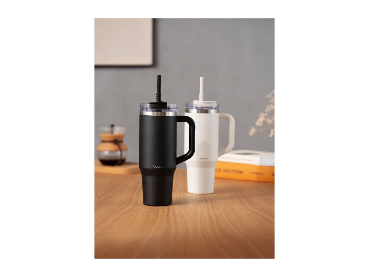Two insulated tumblers with handles and straws on a wooden table, one black and one white.