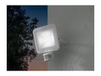 An outdoor LED floodlight with a motion sensor illuminates a textured wall at dusk.