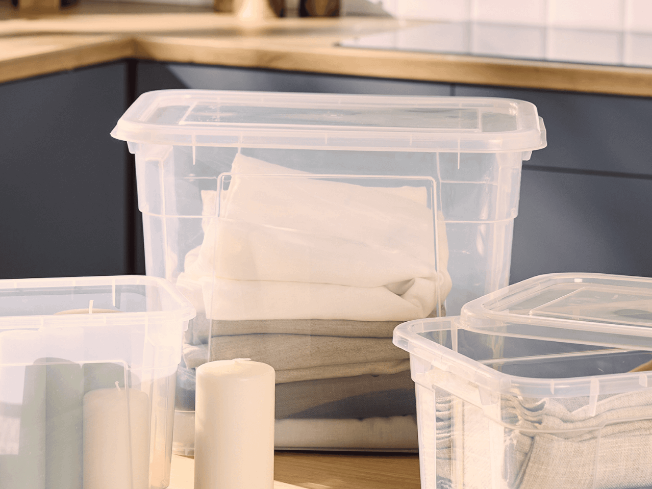 Transparente Aufbewahrungsboxen mit gefalteter Wäsche und Kerzen auf einer Küchenarbeitsplatte.