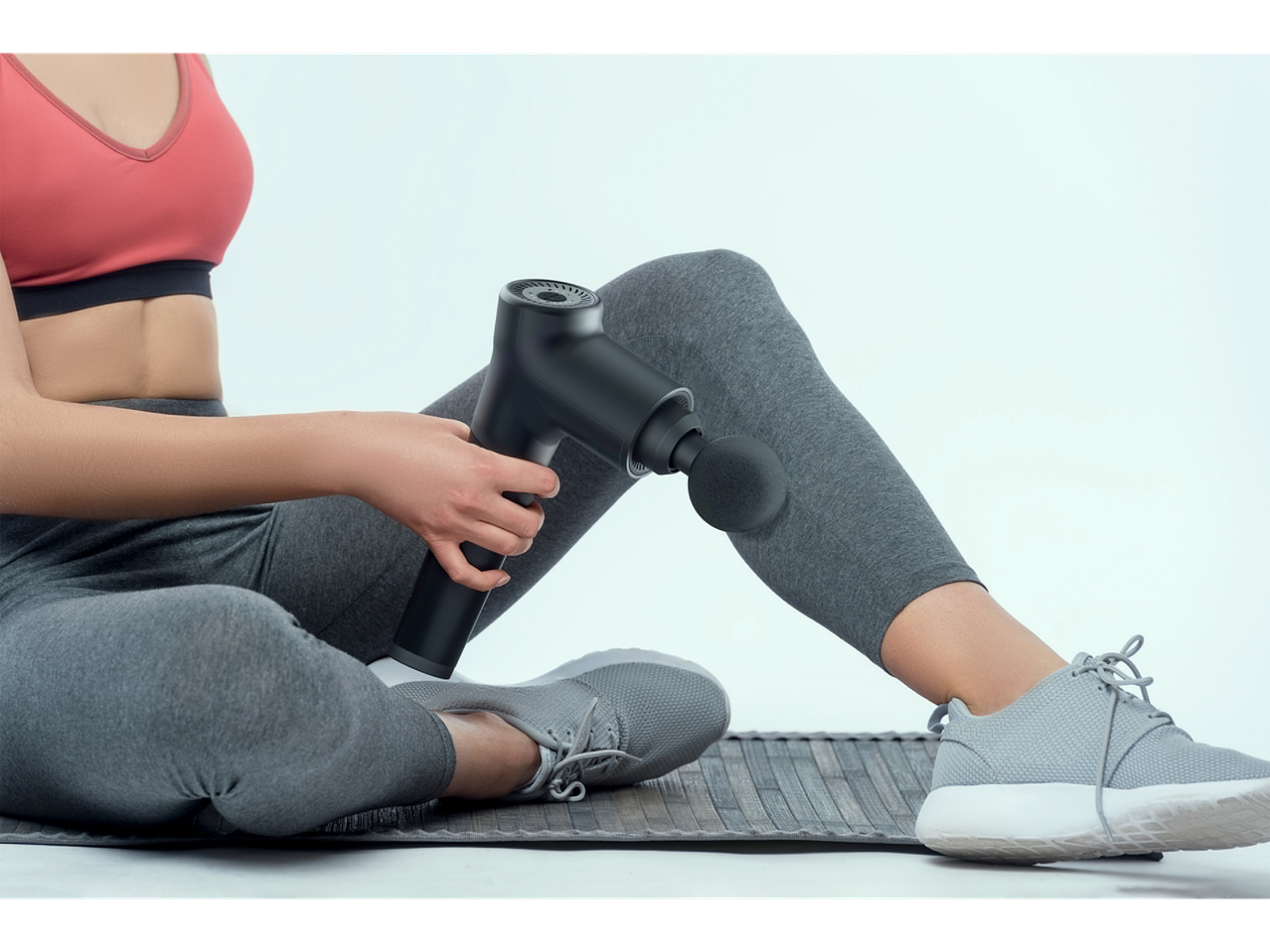 A person in athletic wear uses a black massage gun on their leg while sitting on a mat.