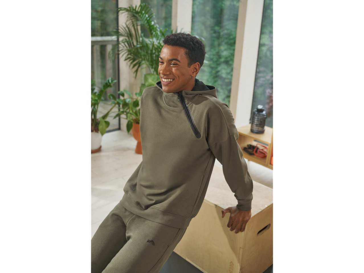 Besišypsantis vyras su CRIVIT funkcionalia striuke ir derančiomis kelnėmis, darantis atsispaudimus ant medinės dėžės.