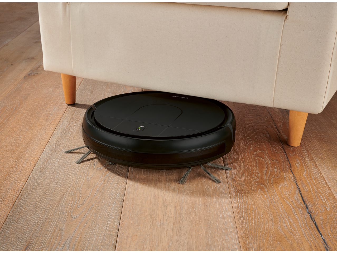 Crni robotski usisivač čisti ispod krem sofe na drvenom podu.