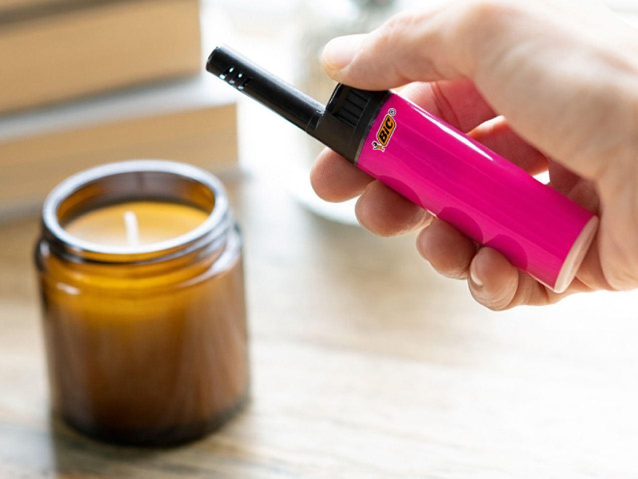 A hand holds a pink long lighter to light a candle in an amber jar.