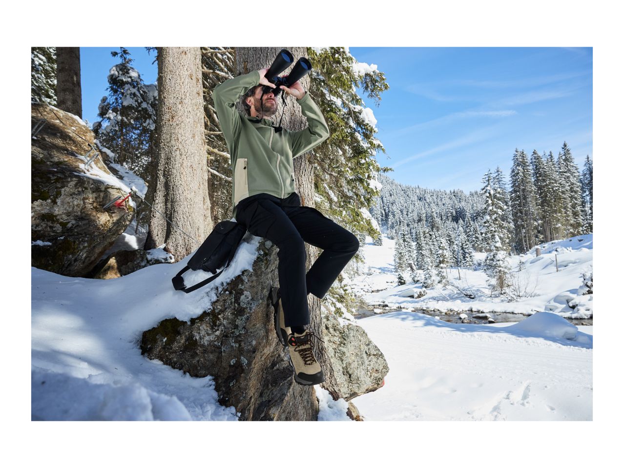 A man in a green outdoor jacket and black trousers looking through binoculars in a snowy forest.