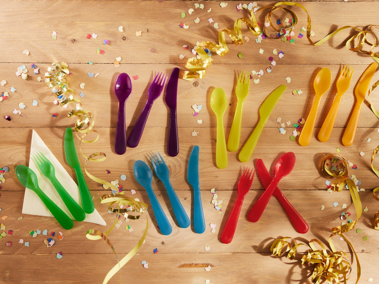 Разноцветни пластмасови прибори за хранене и конфети върху дървена маса.