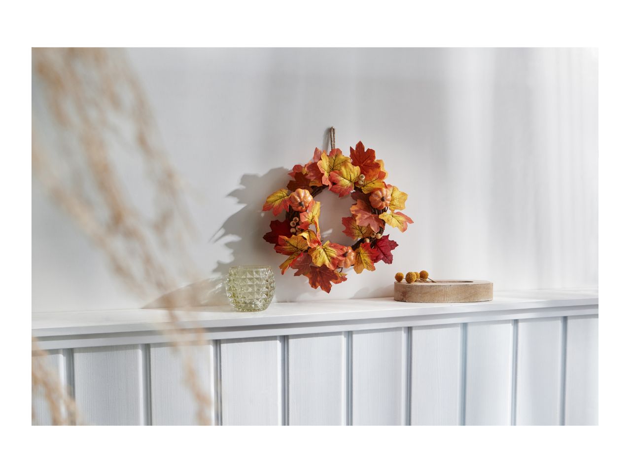 Autumn wreath with pumpkins and leaves, a glass candle holder, and a wooden tray.