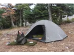 A woman in a sleeping bag sits outside a tent with camping gear in a forest.