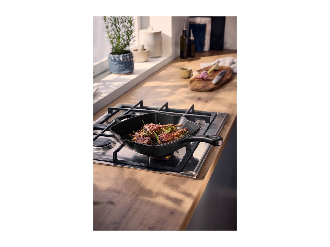 Cast iron grill pan with meat skewers on a gas hob in a kitchen setting.