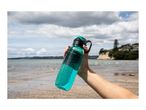 A hand holding a teal reusable water bottle at a beach.