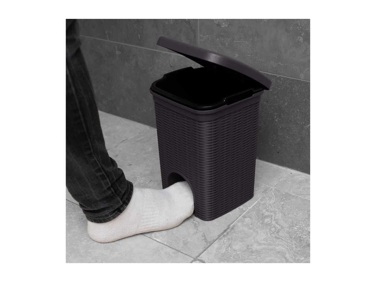 Foot operating a dark grey pedal bin with a woven texture, lid open, on a tiled floor.