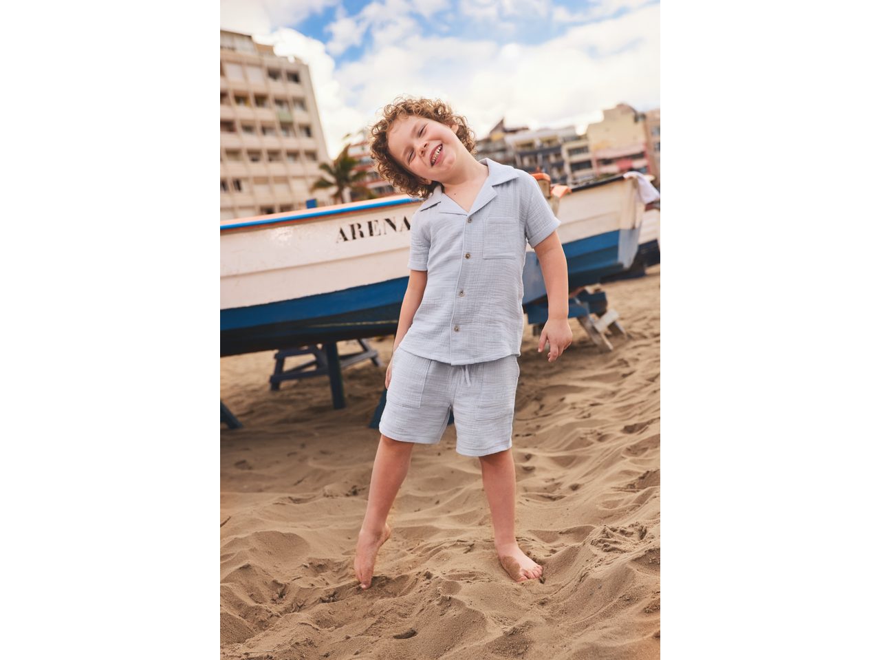 Smiling boy in a light blue short-sleeved shirt and shorts, standing barefoot on a sandy beach.