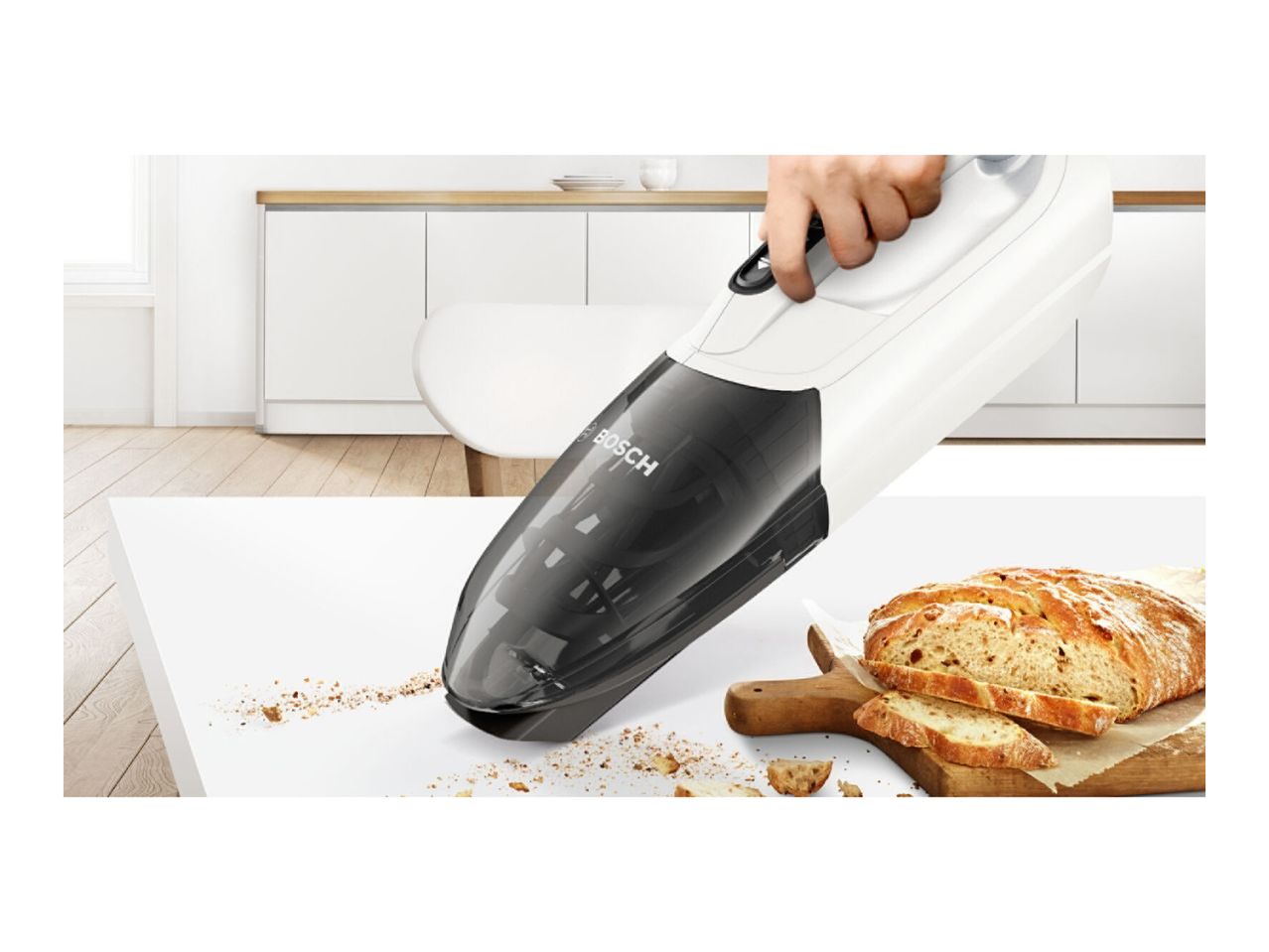 A Bosch hand-held vacuum cleaner cleans crumbs from a kitchen table near a loaf of bread.
