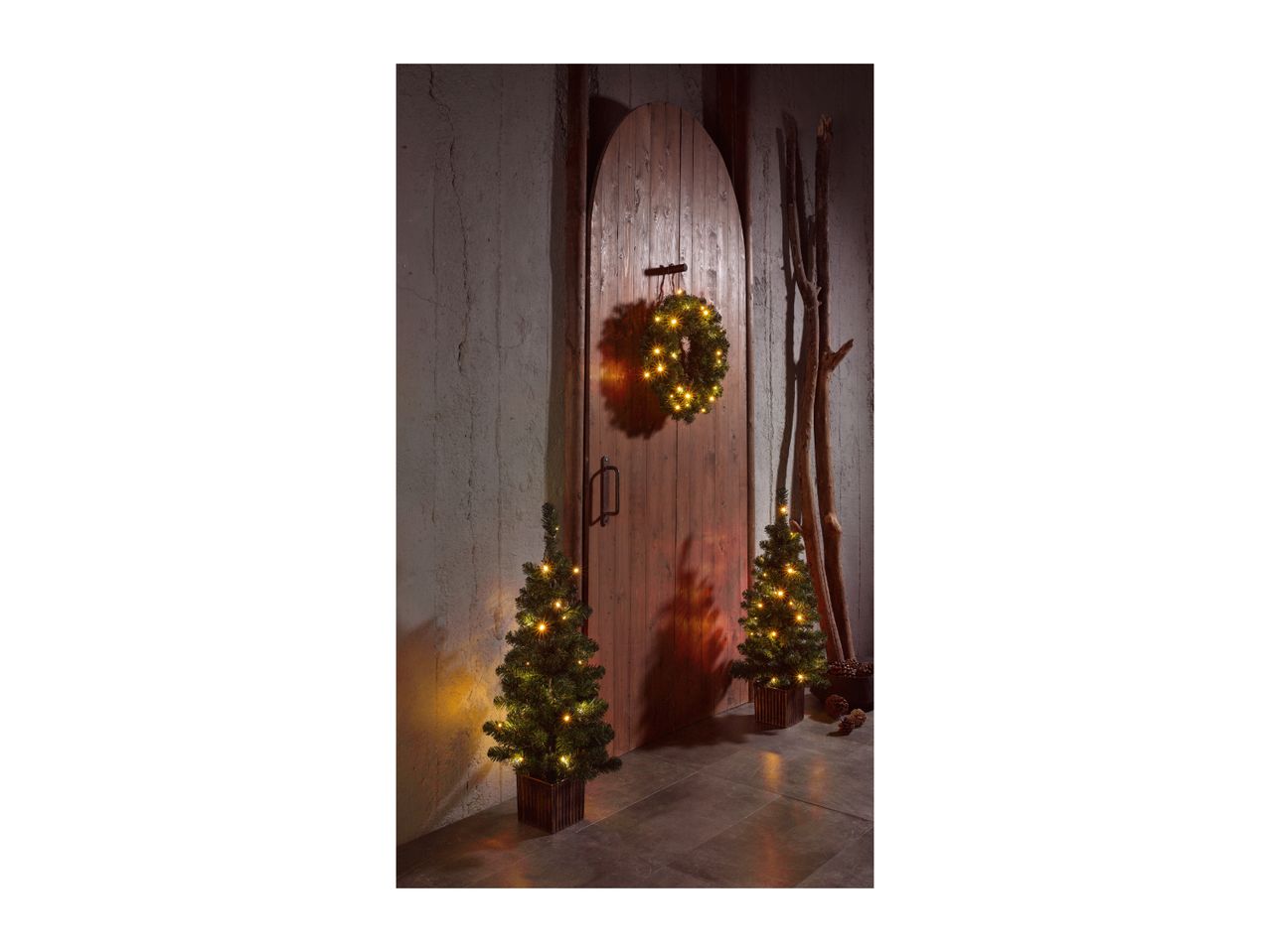 Two small Christmas trees and a wreath with warm LED lights, set against a rustic wooden door.