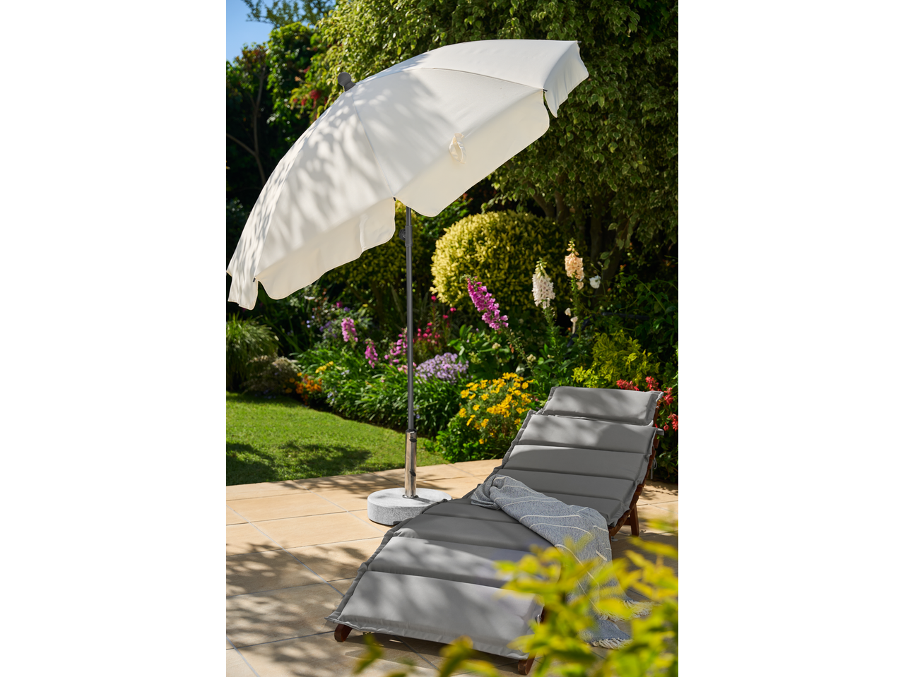 Guarda-sol LIVARNO branco num pátio com espreguiçadeira cinzenta e jardim florido.