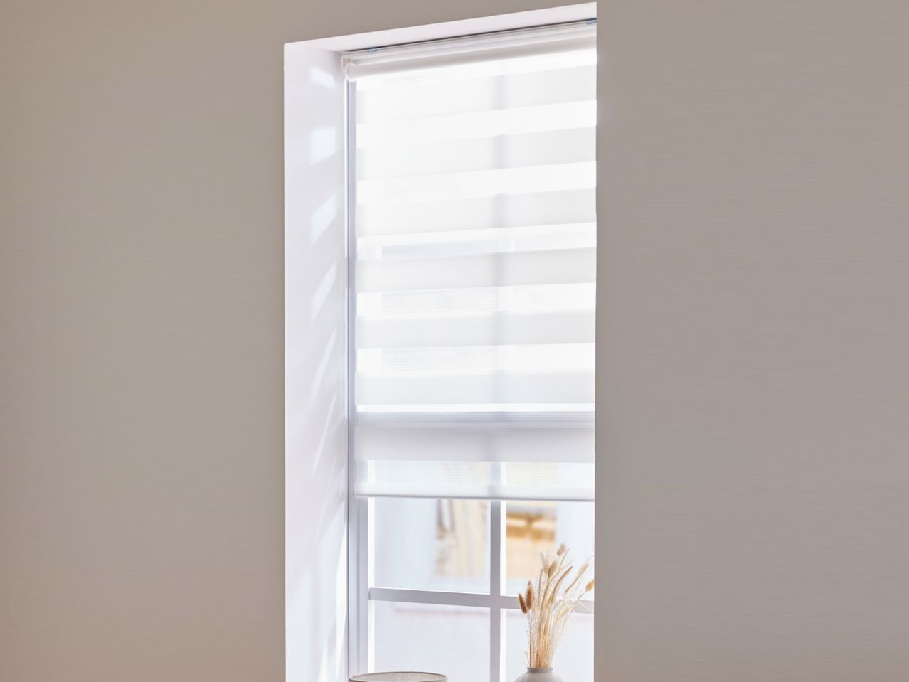 Prozor s bijelim roletama i vazom sa suhim biljem na prozorskoj dasci.