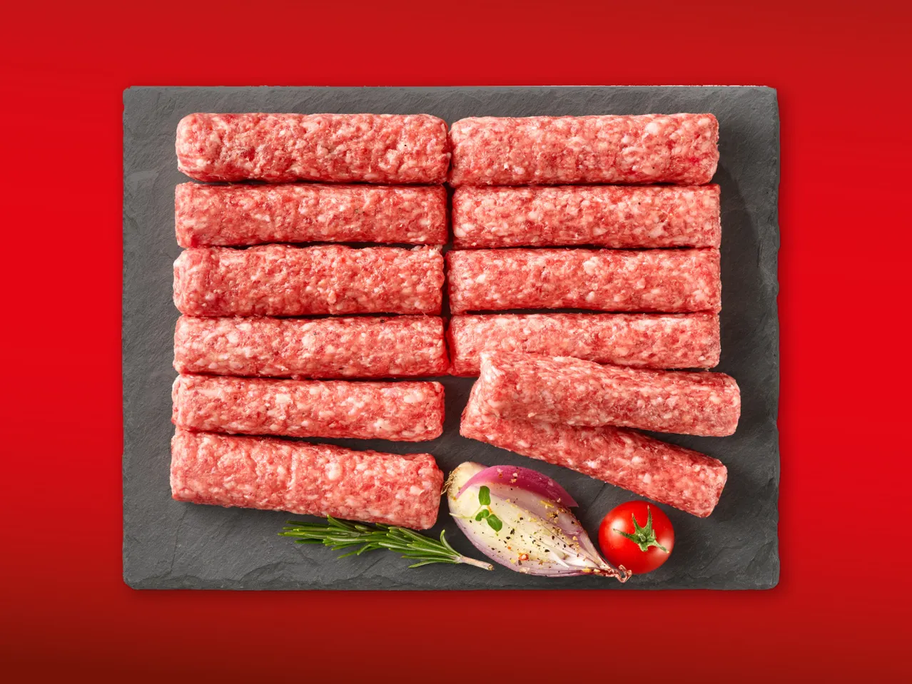 Sirovi ćevapi na crnoj ploči s rajčicom, lukom i ružmarinom.
