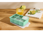 Forma de gelo verde menta com cubos de gelo grandes, uma bebida e rodelas de lima numa mesa de madeira.
