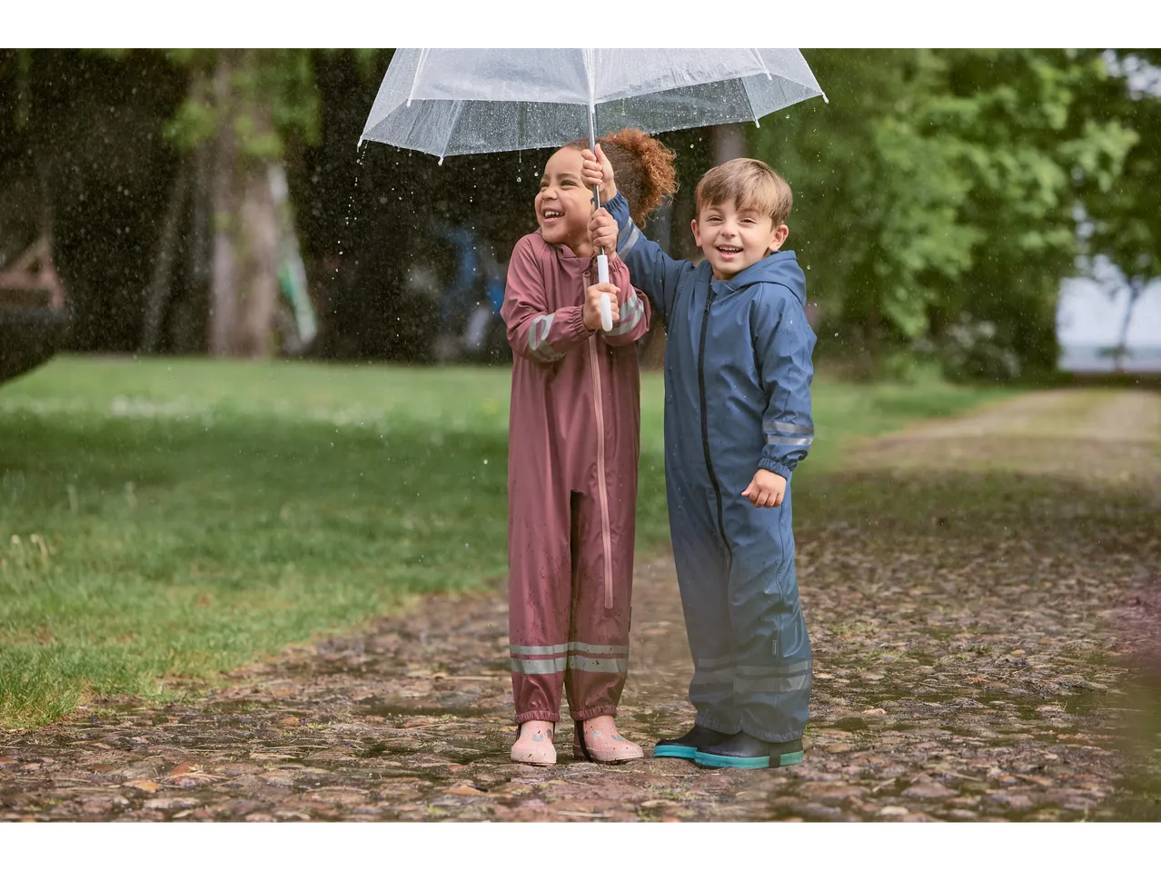 Doi copii în costume de ploaie și cizme sub o umbrelă, în ploaie.