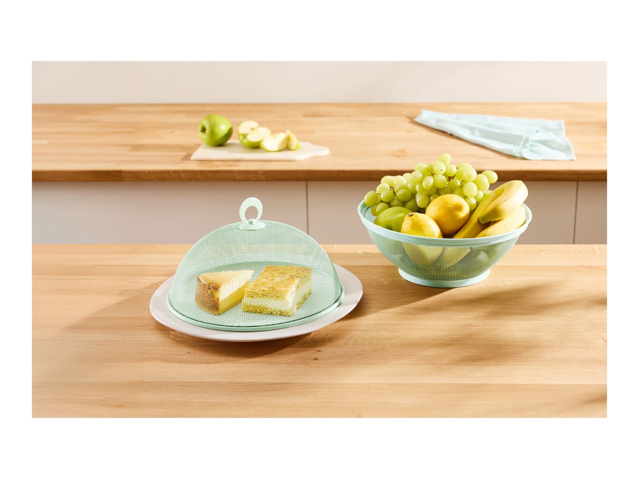 Kitchen counter with cake under a mesh cover, fruit bowl, and chopped apples.